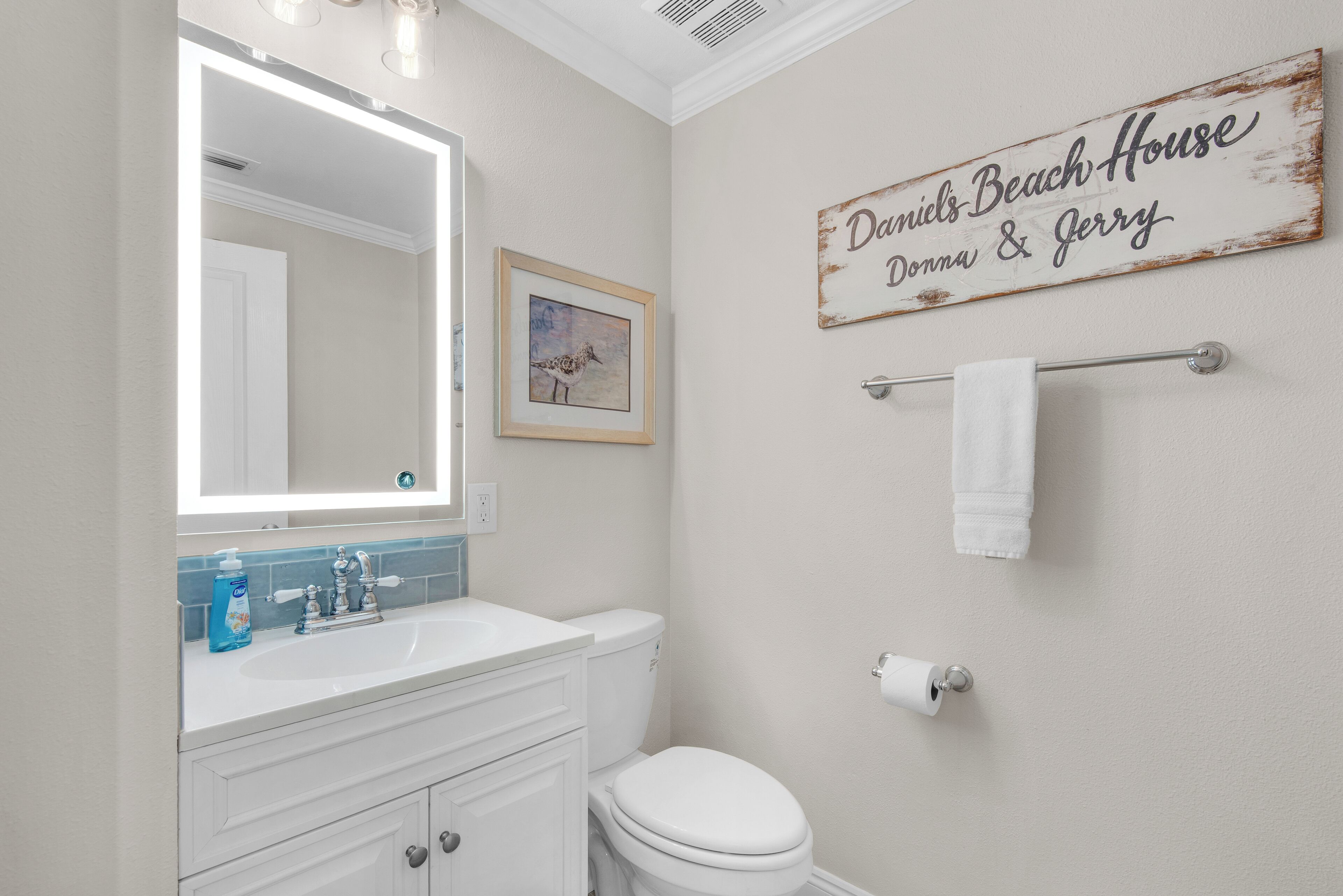 A bathroom with a white sink, mirror, and toilet. There is a framed picture on the wall and a sign that reads "Daniel's Beach House Donna & Jerry".