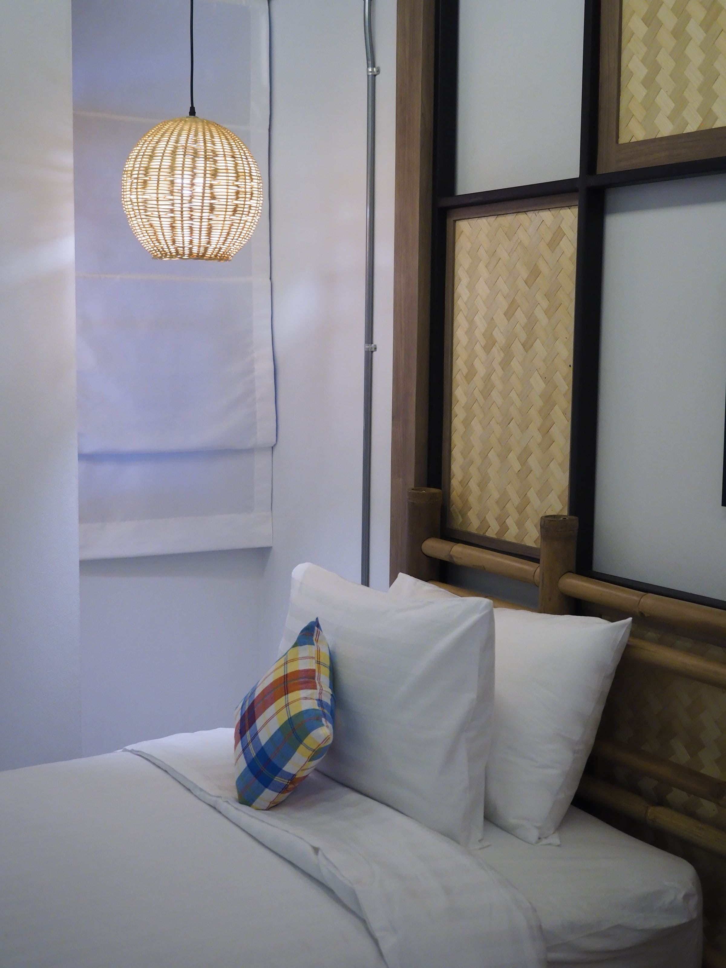 A neatly made bed with white linens and a colorful pillow, a woven pendant light, and a wooden headboard with a woven back panel.