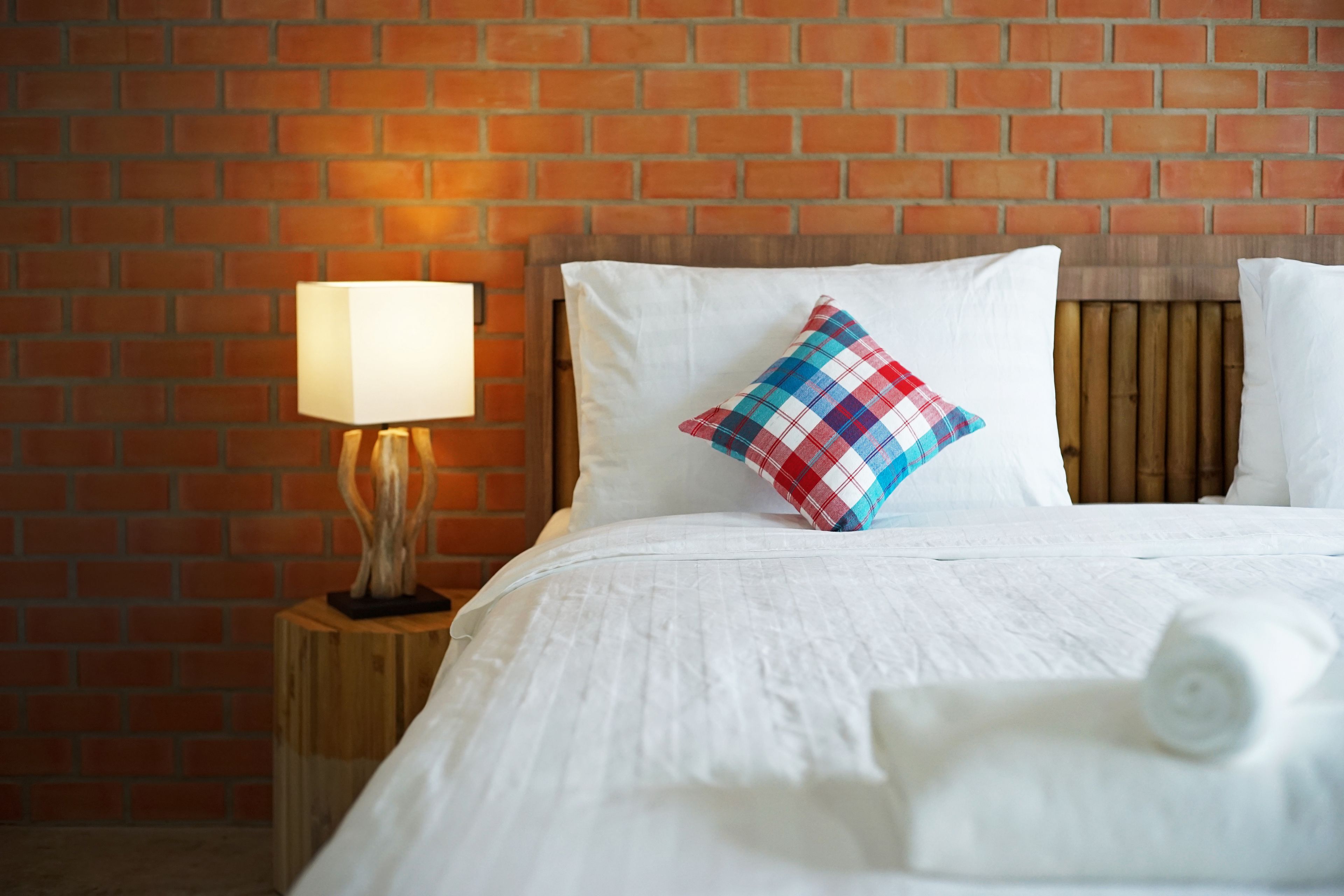 A neatly made bed with white linens, a plaid pillow, and a rolled towel on a wooden nightstand with a lamp.