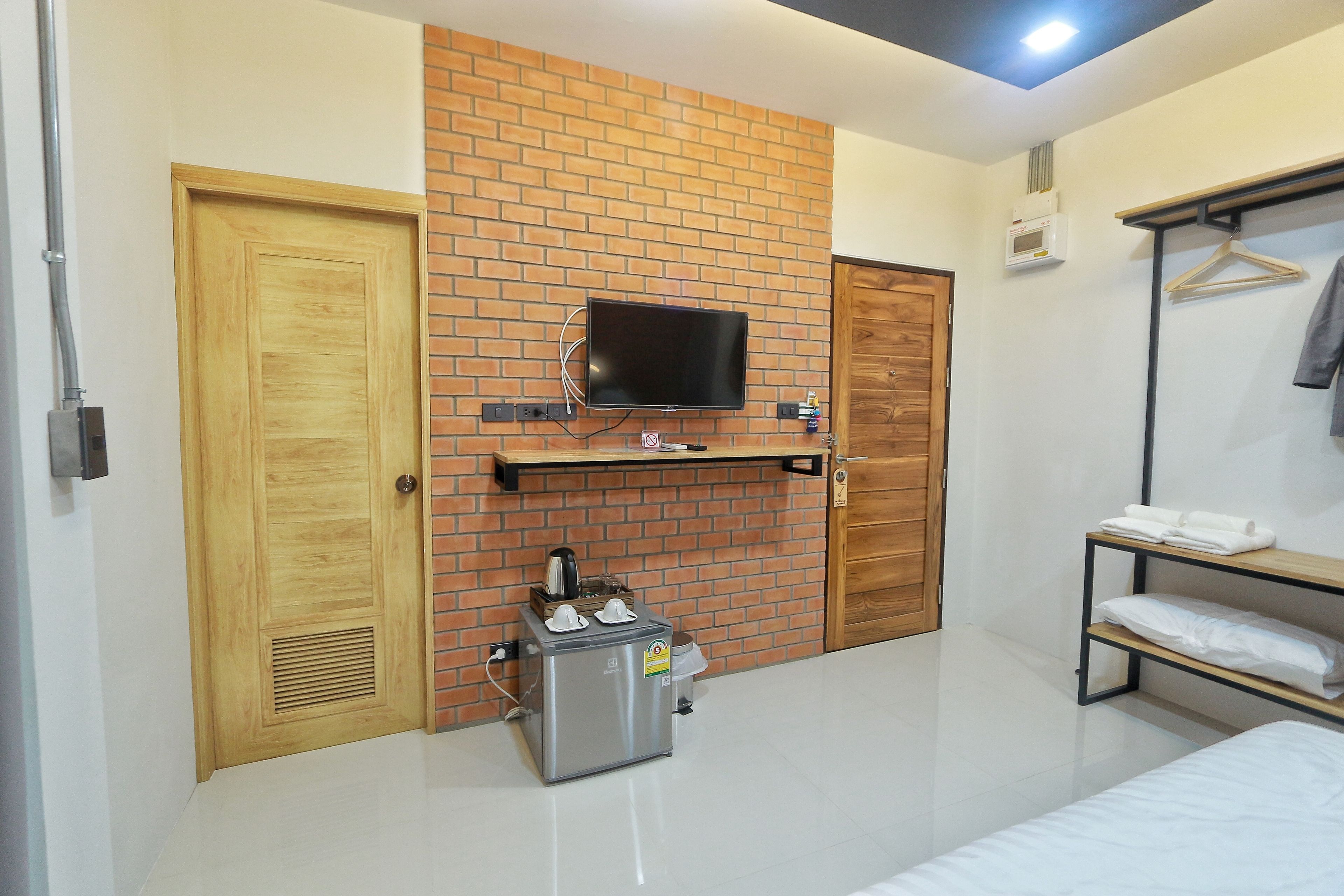 Une chambre d’hôtel moderne avec un mur d’accent en briques, un téléviseur à écran plat fixé au-dessus, une petite kitchenette, un lit, une porte en bois et un portant à vêtements.