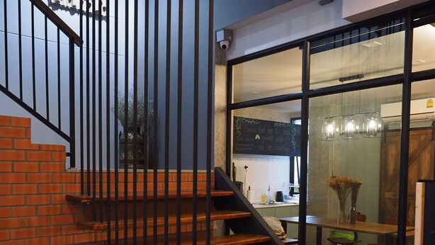 A staircase with wooden steps and black metal railings, leading to a room with a dining area visible through a glass door. - Luck Esan Loft - Hostel (Ubon Ratchathani)