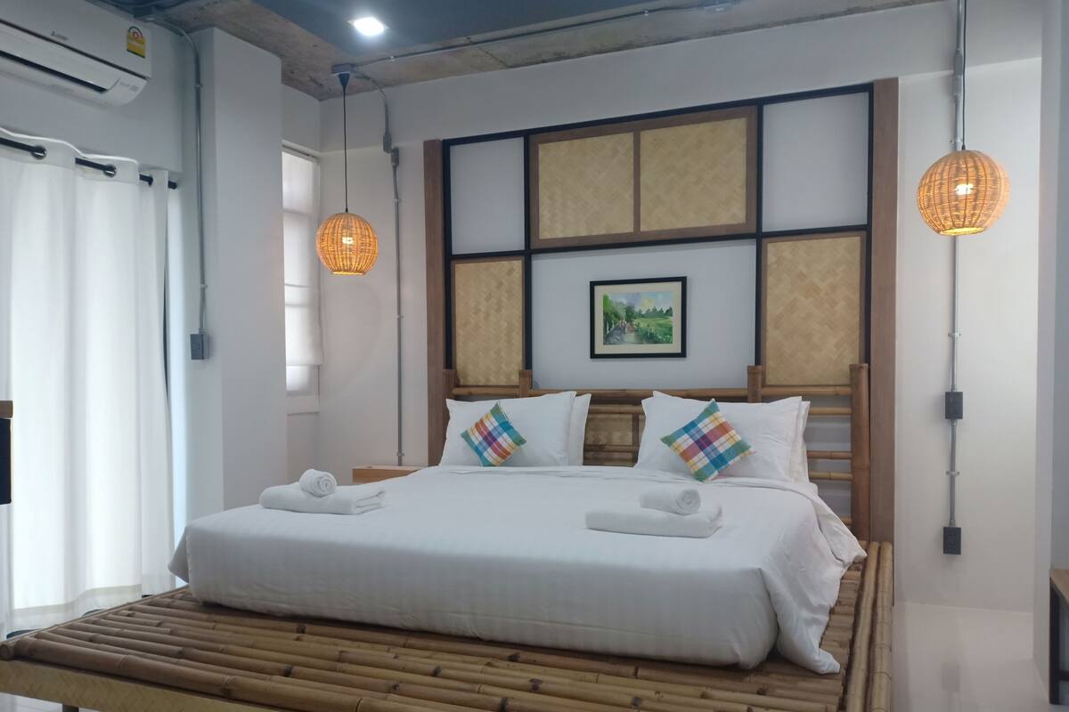 A neatly made bed with white linens and two colorful pillows, a wooden headboard, and a framed picture on the wall.