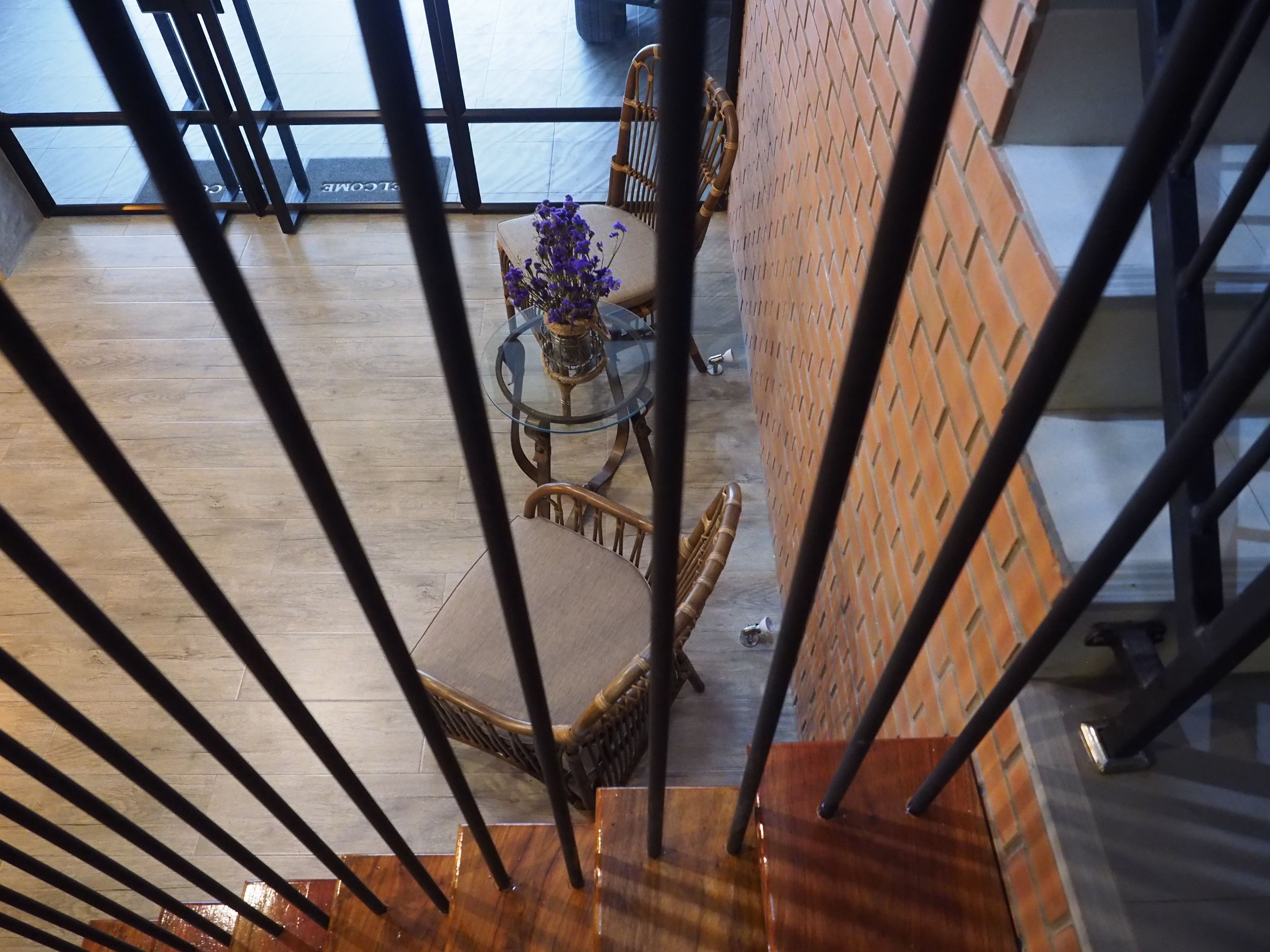 Een modern interieur met een houten trap, een glazen tafel met paarse bloemen, rieten stoelen en een bakstenen muur.