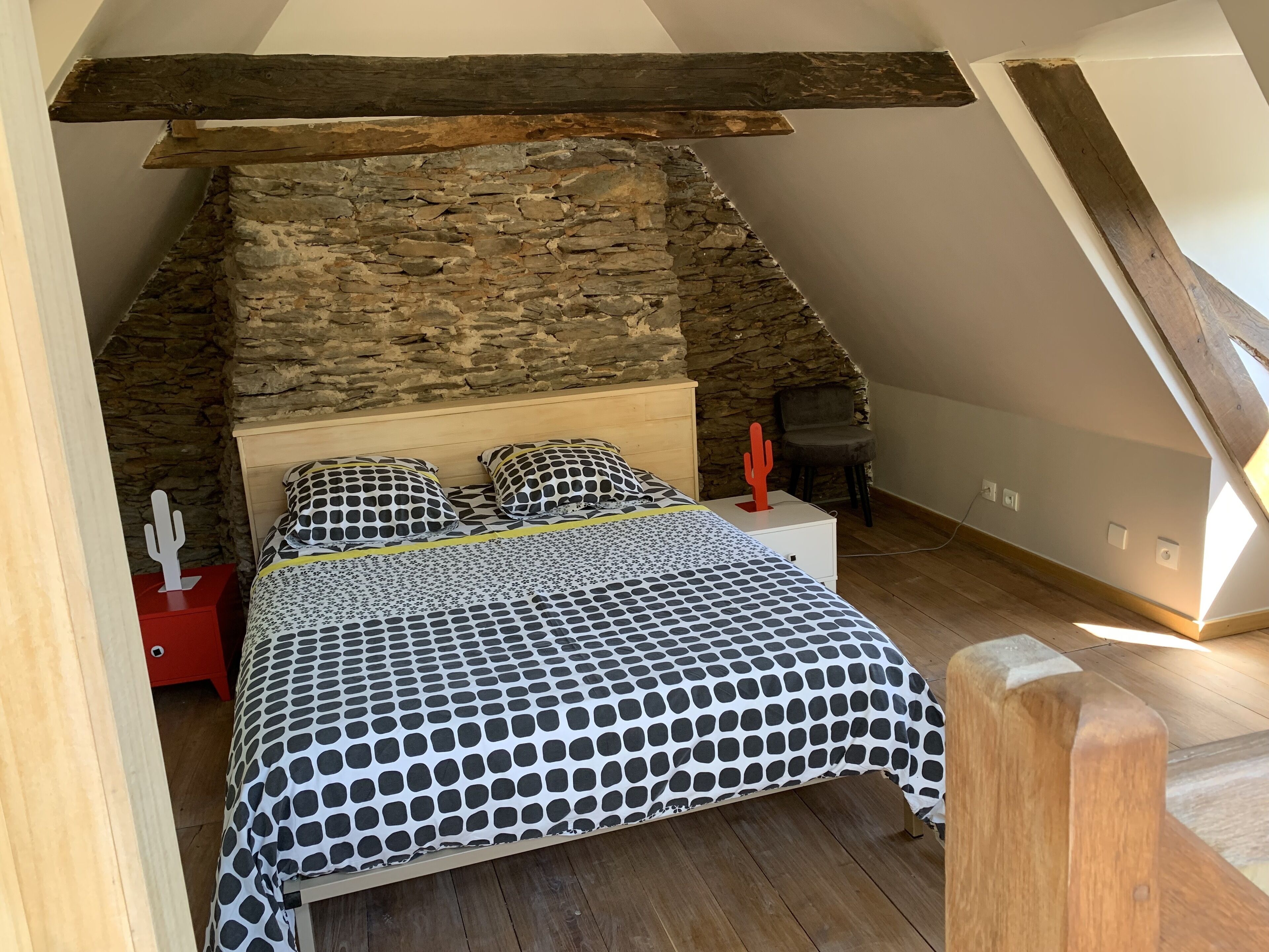 Un dormitorio con pared de piedra, vigas de madera, una cama con colcha de lunares, una decoración de cactus y una silla.