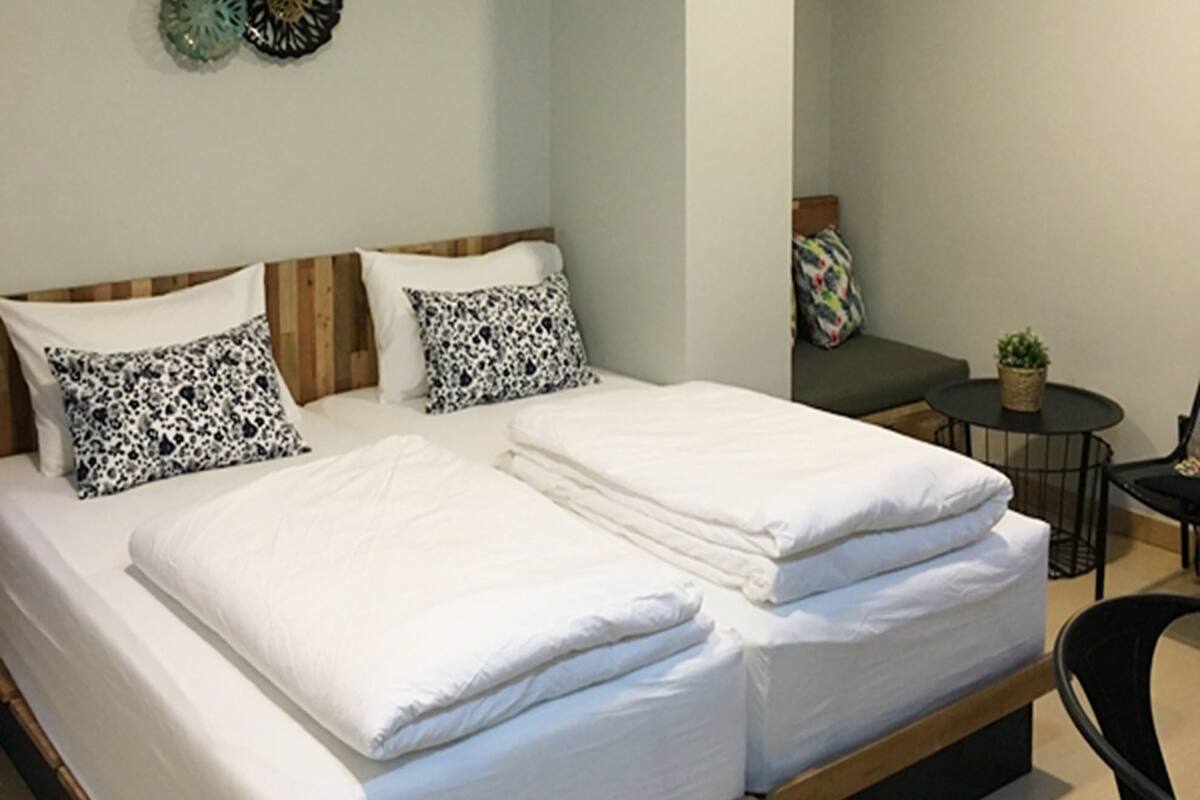 A neatly made double bed with white linens, a small round table with a plant, and a chair in a bedroom with light-colored walls and a decorative wall piece.