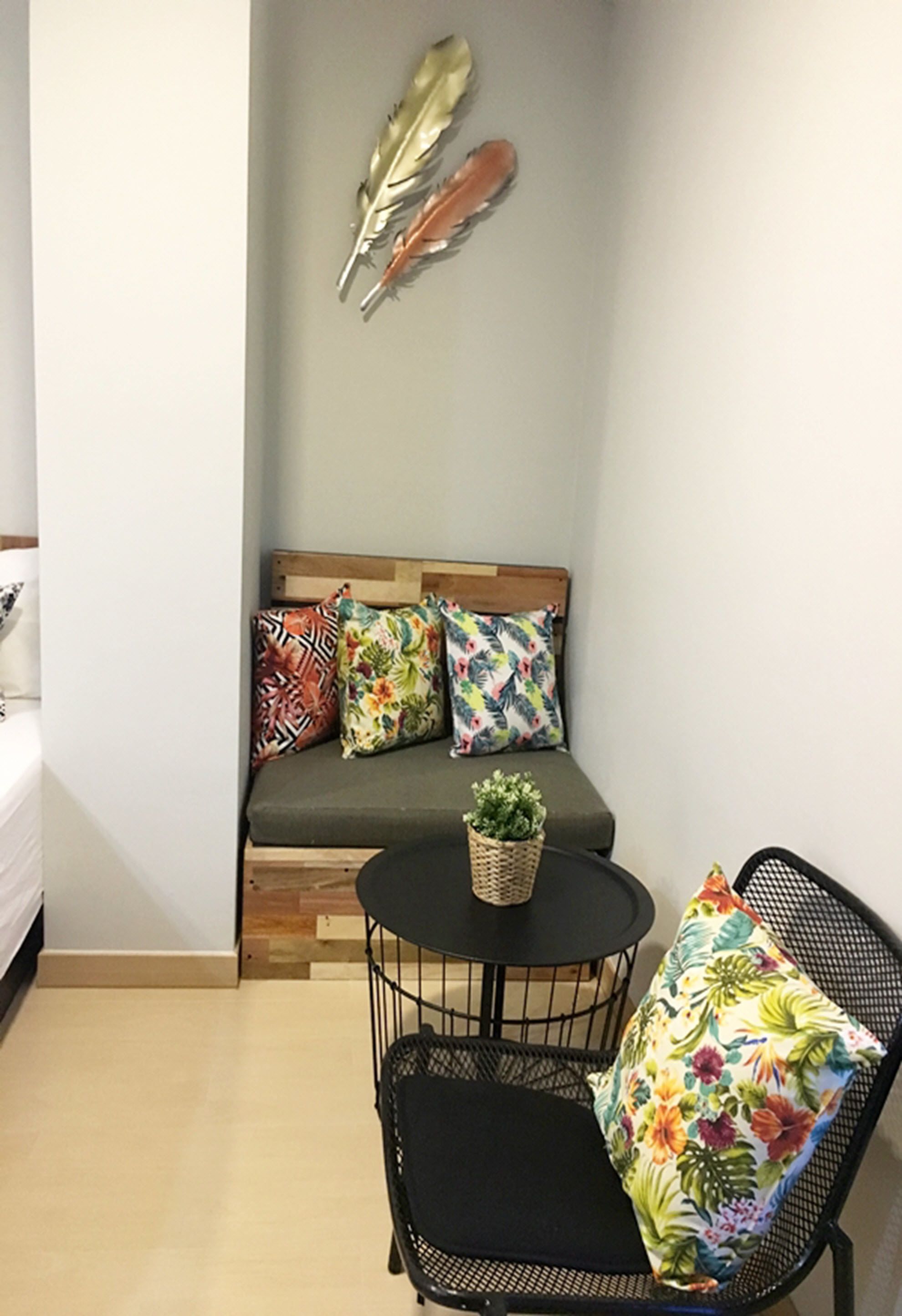 Una habitación pequeña con una cama, un sofá con almohadones de colores, una mesita con una planta y una silla con un cojín estampado.