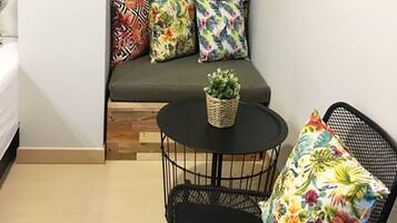 Una habitación pequeña con una cama, un sofá con almohadones de colores, una mesita con una planta y una silla con un cojín estampado.