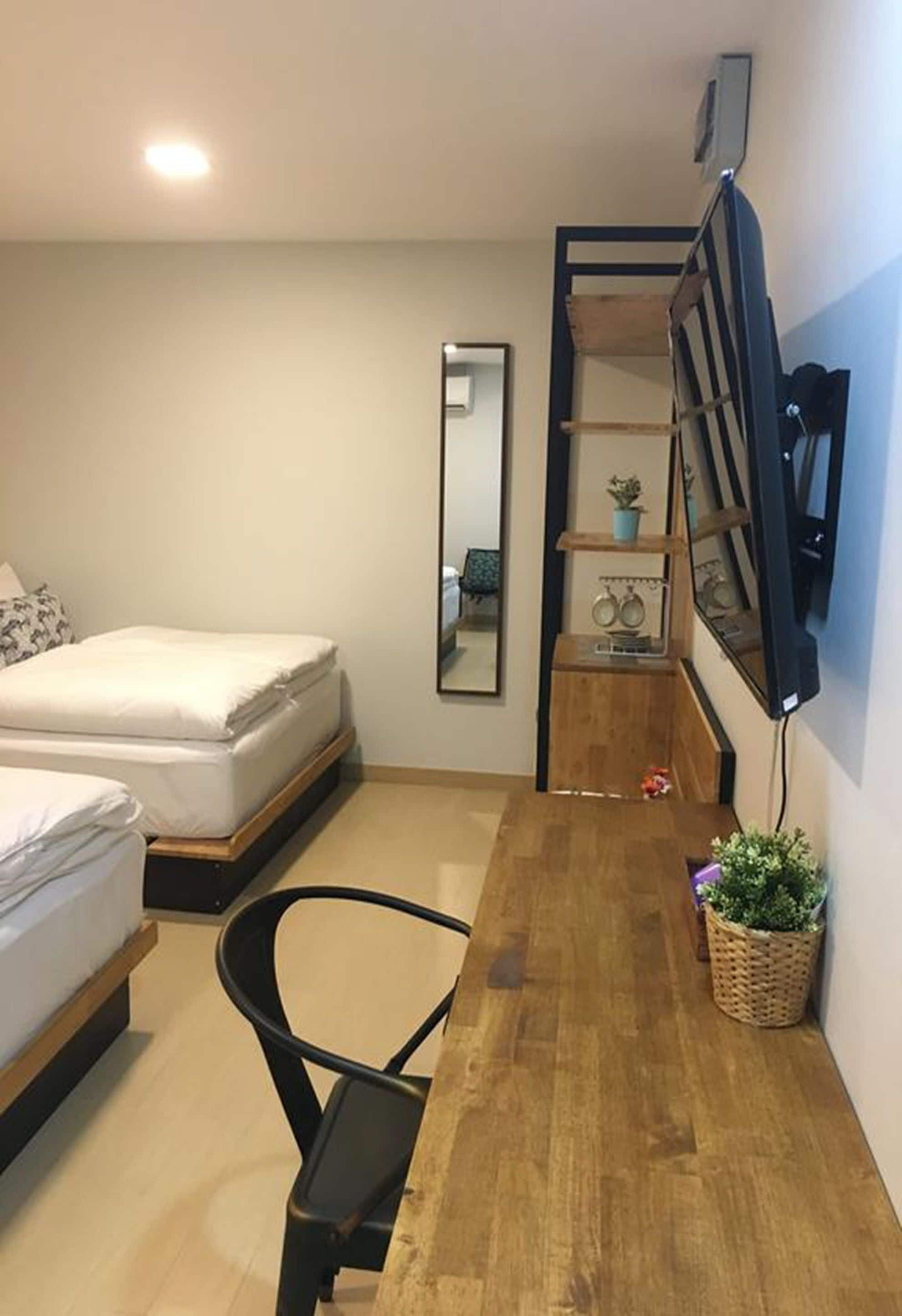 A small, well-lit room with a bed, a desk, a chair, a TV mounted on the wall, and a mirror.
