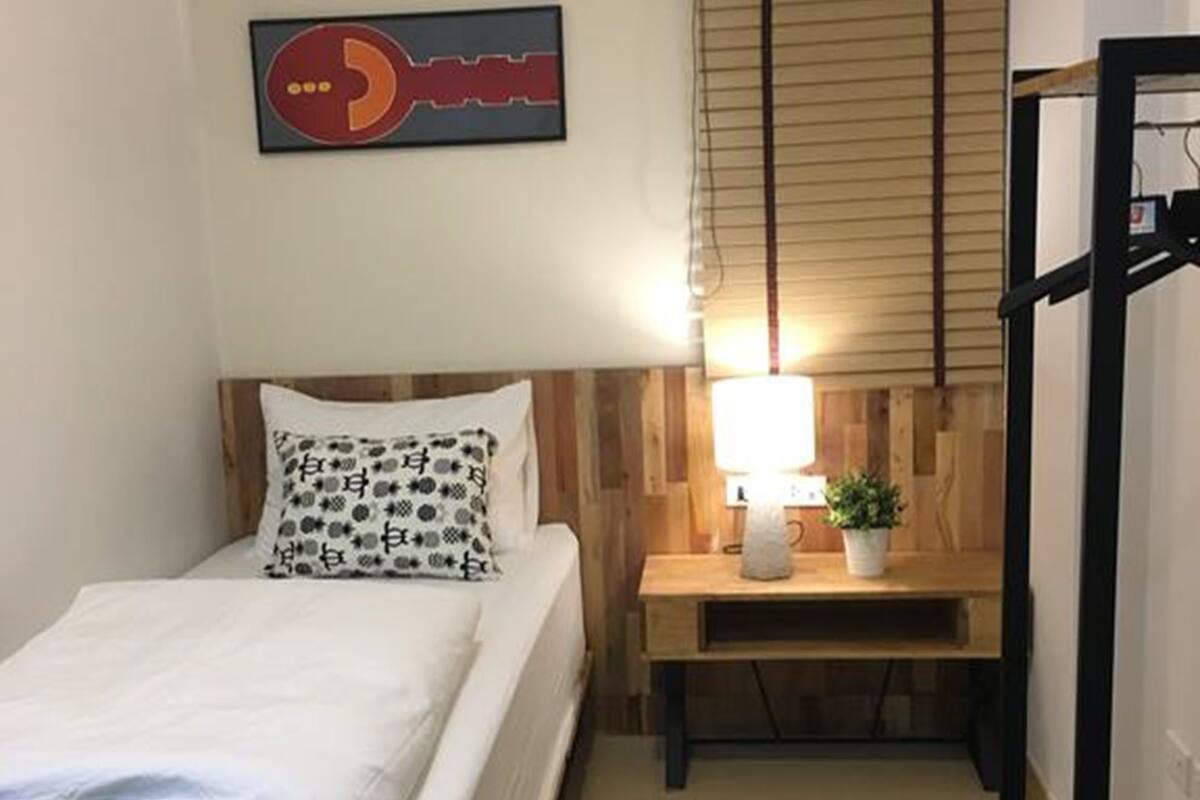 A small, neatly arranged room with a bed, bedside table, lamp, and air conditioning unit.