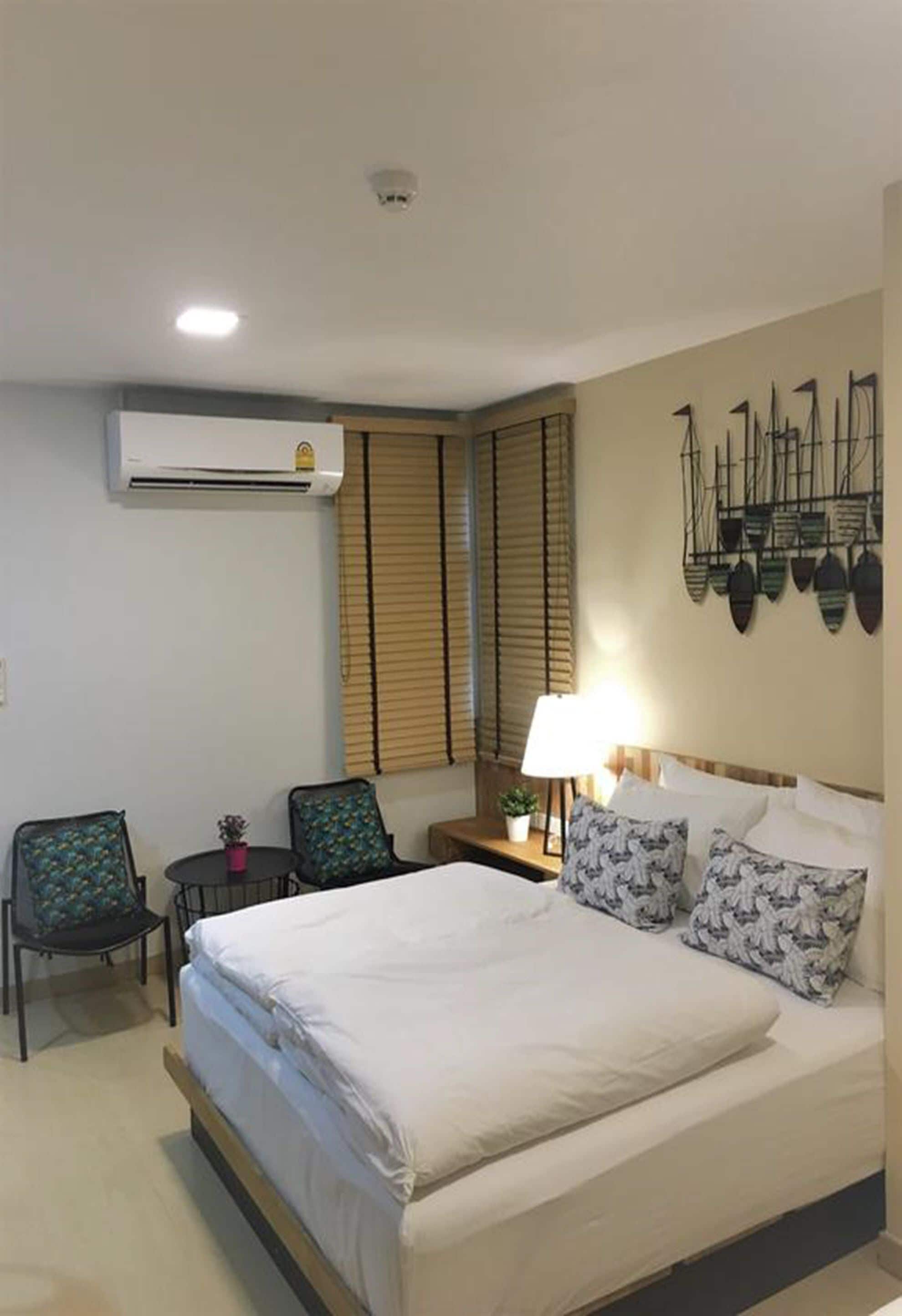 Habitación de hotel con cama, dos sillas, una mesita de noche, lámpara y aire acondicionado instalado en la pared.