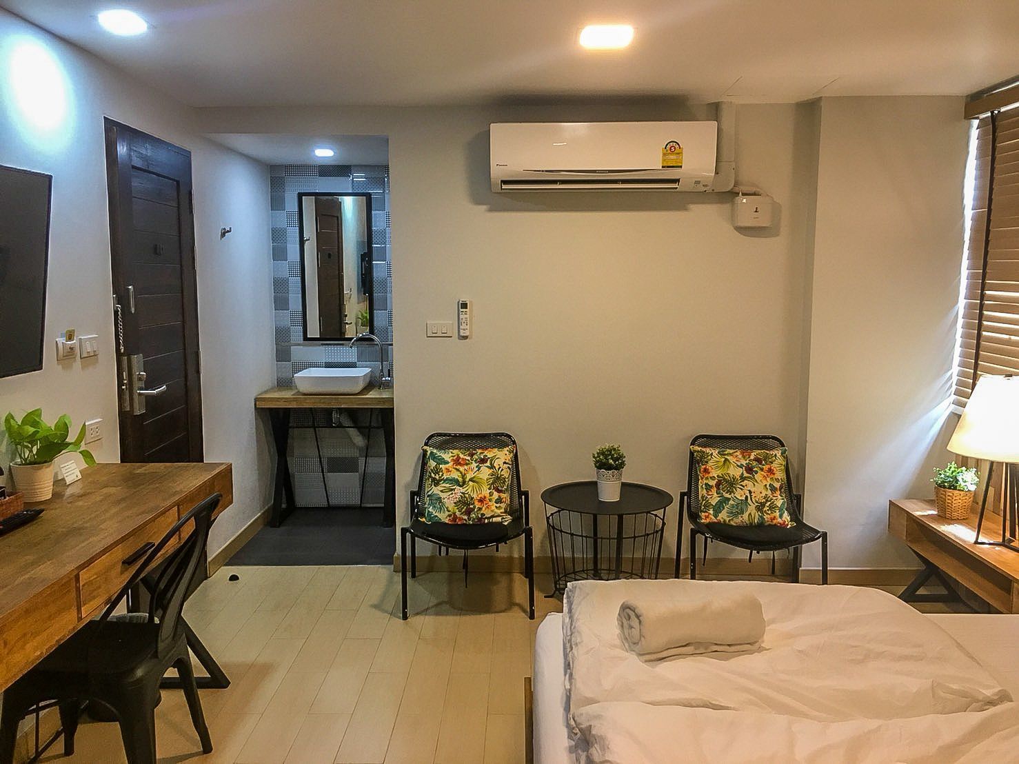 A compact hotel room with a bed, a desk with a chair, a small table with two chairs, a bathroom with a sink and mirror, and an air conditioning unit.
