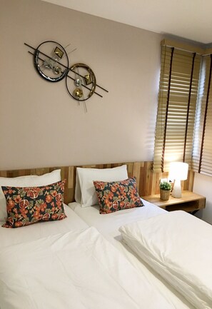 Habitación de hotel con cabecera de madera, dos camas blancas, almohadas estampadas con motivos florales, un reloj de pared y una lámpara de noche.