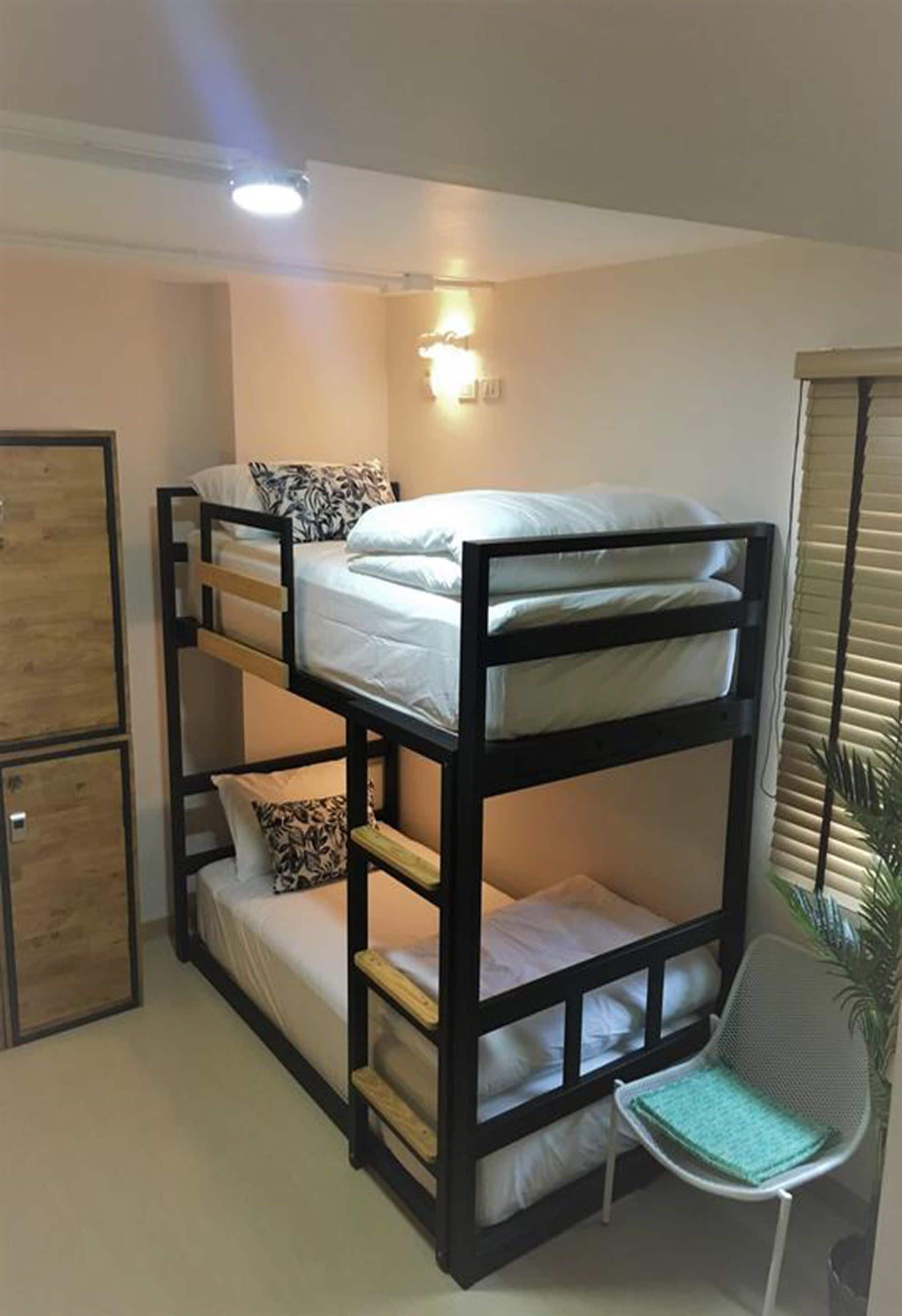 Une chambre avec un lit superposé, un bureau et une chaise, un petit placard et une fenêtre avec des stores.