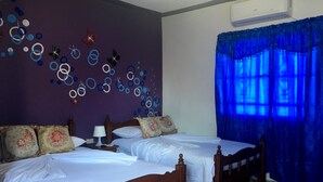 Ein Zimmer mit zwei Holzbett, einer Wand mit kreis- und floralen Verzierungen, einem Fenster mit blauen Vorhängen und einer Klimaanlage.