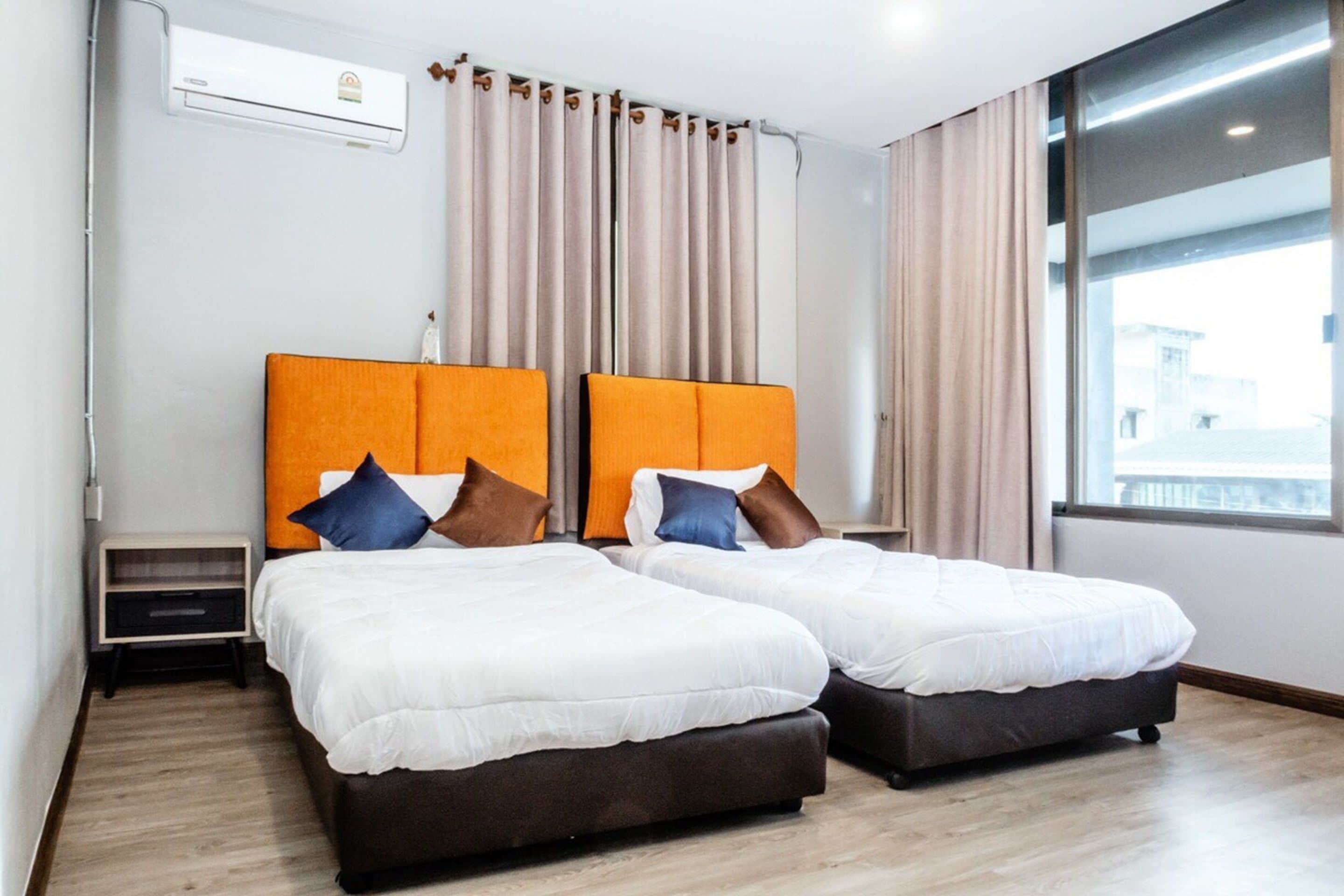 A hotel room with two beds, a window with curtains, an air conditioning unit, and a small bedside table.
