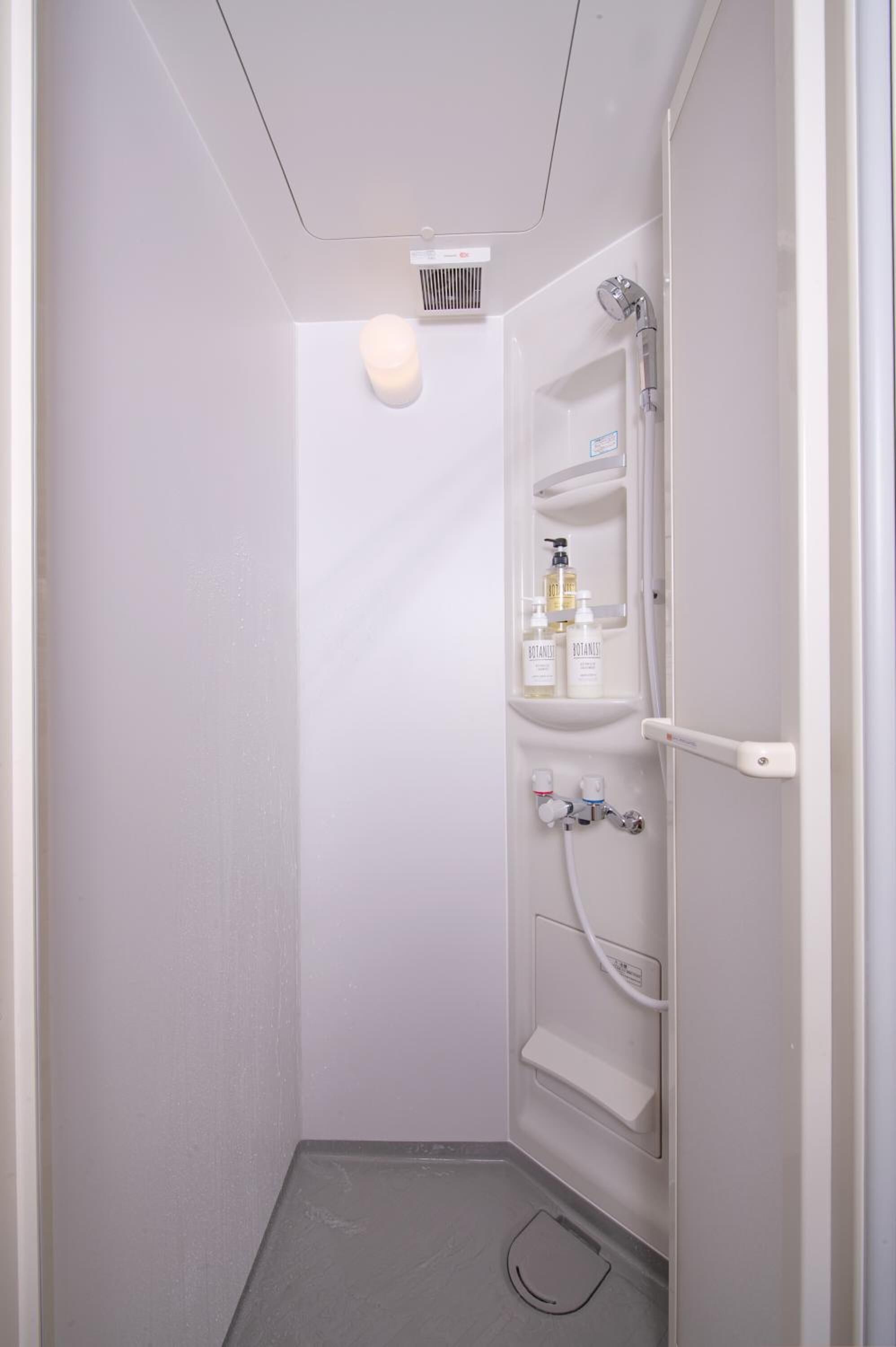 A small, white bathroom with a shower, a toilet, and a shelf with toiletries.