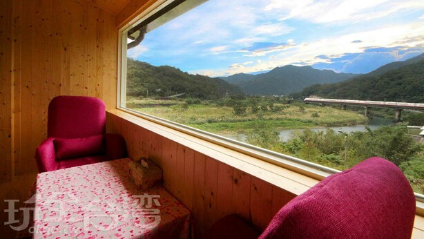 Una cabaña de madera con una mesa y dos sillas, una ventana que ofrece vistas a un río y montañas, y un tren en un puente a lo lejos.