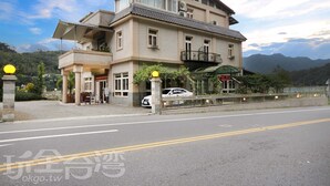 Un hotel de dos pisos con balcón, entrada cubierta y un coche estacionado frente a él.
