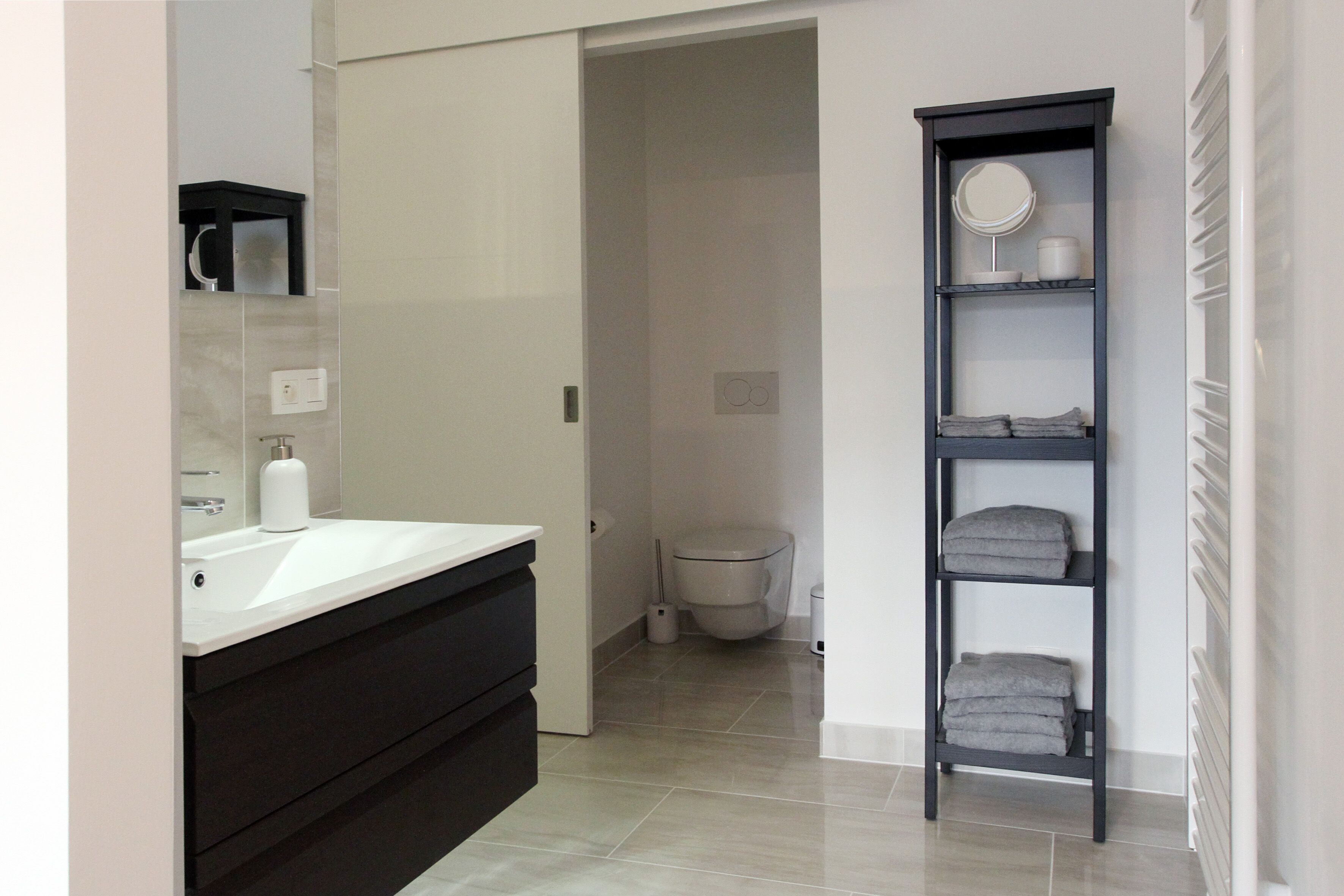 Une salle de bain moderne avec un lavabo blanc, des meubles sombres, une haute étagère noire où sont posées des serviettes, et des toilettes à l’arrière-plan.