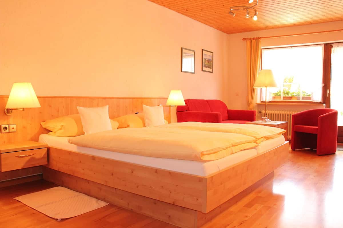 Quarto de hotel com cama de madeira, duas luminárias de cabeceira, uma cadeira vermelha e uma janela com cortinas.