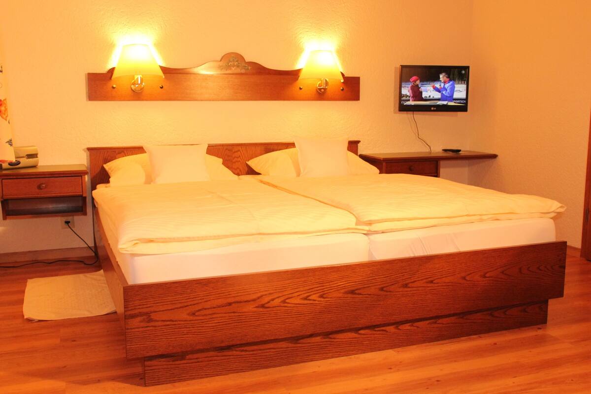 Uma cama de casal com cabeceira e peseira de madeira, duas mesas de cabeceira, uma televisão fixada na parede e uma mesa de apoio de madeira com uma lâmpada.