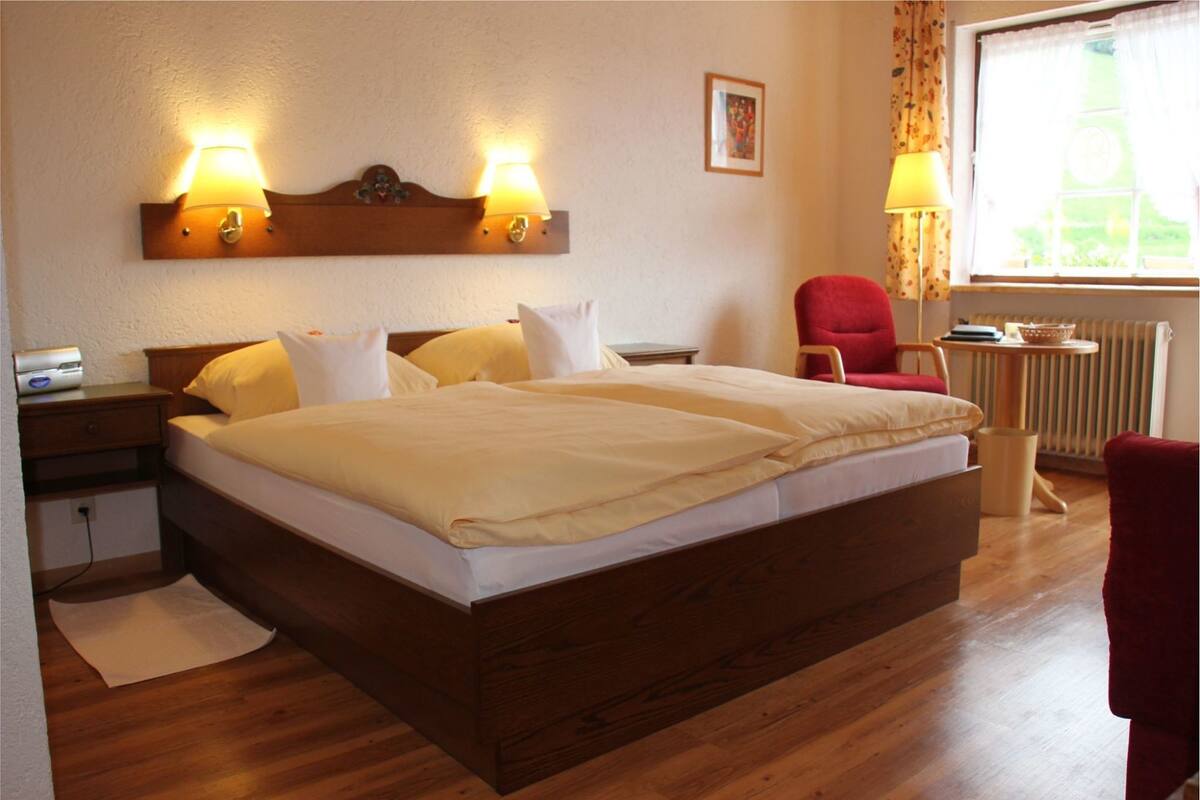 Cama bem arrumada com duas luminárias de cabeceira, uma cadeira vermelha e uma janela com cortinas.