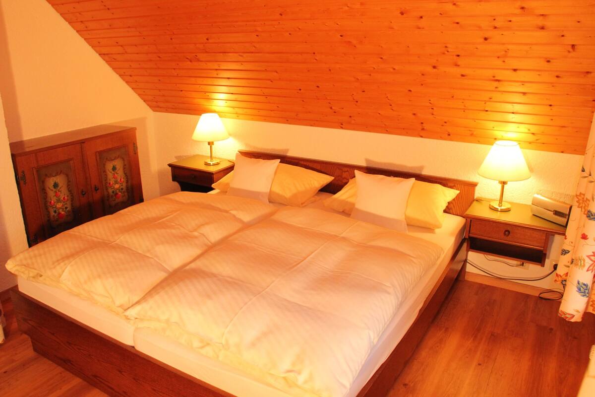 Quarto com teto de madeira, uma cama grande com roupa de cama branca, duas mesas de cabeceira com abajures e um guarda-roupa de madeira.