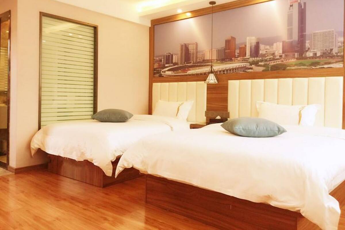 Habitación de hotel con dos camas, un ventanal con persianas, un mural con vista a la ciudad y suelo de madera.