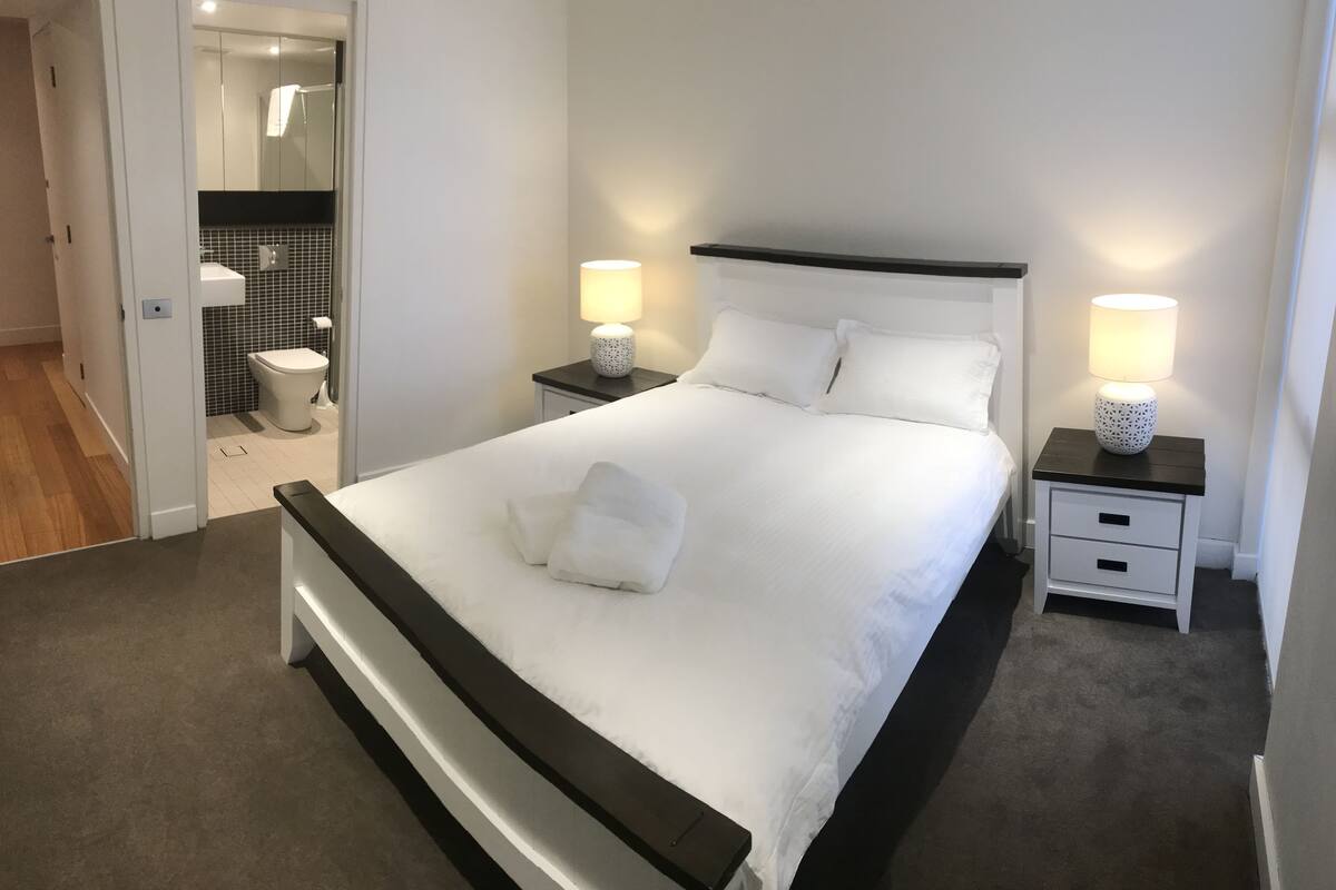 Ein ordentlich bezogenes Bett mit weißen Leinentüchern und einem gefalteten Handtuch, zwei Nachttische mit Lampen, ein dunkles Kopfteil und Blick in ein Badezimmer mit Toilette und Waschbecken.