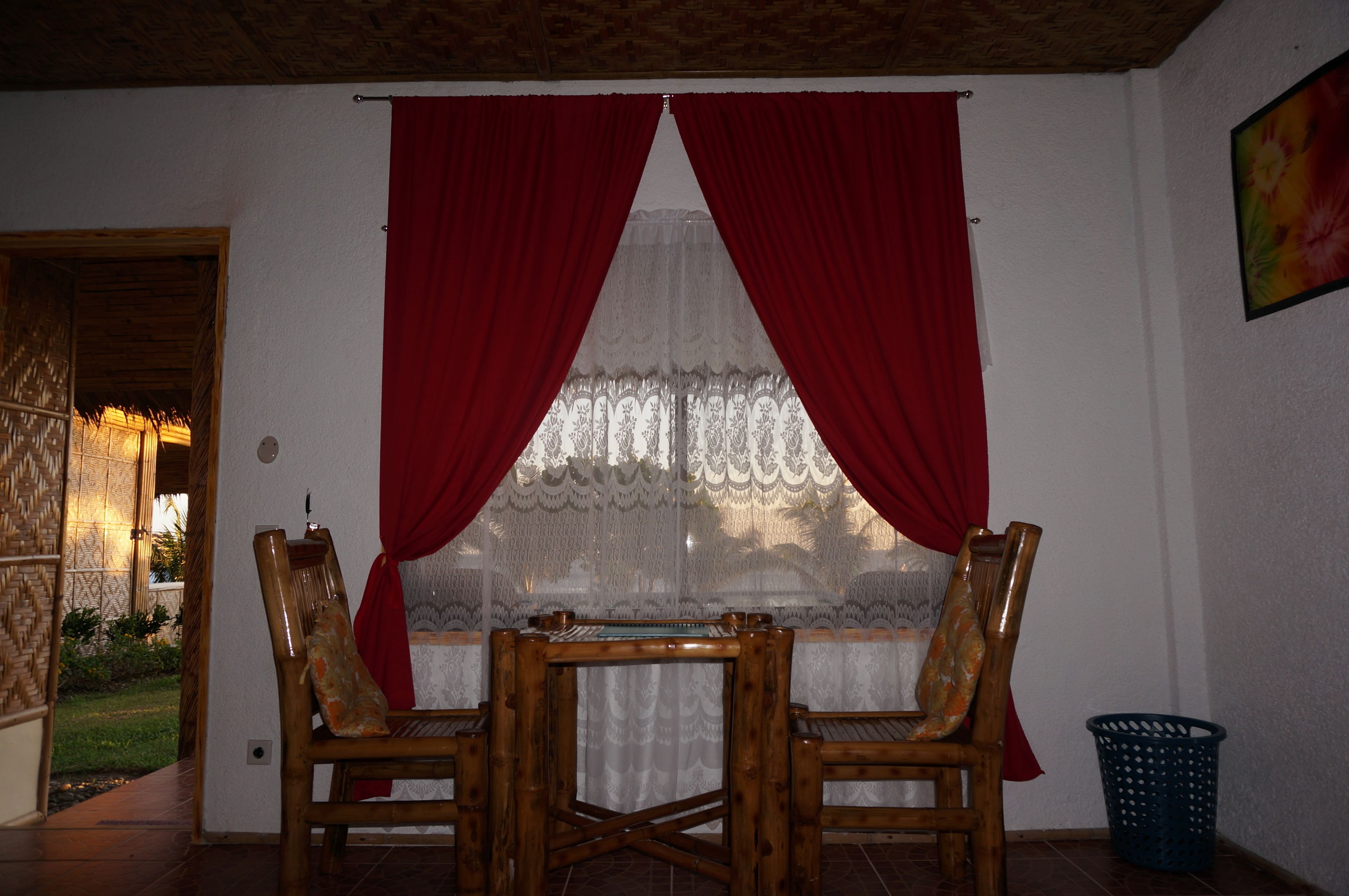 Una habitación con una mesa y sillas de madera, una ventana con cortinas rojas y un papel tapiz estampado.