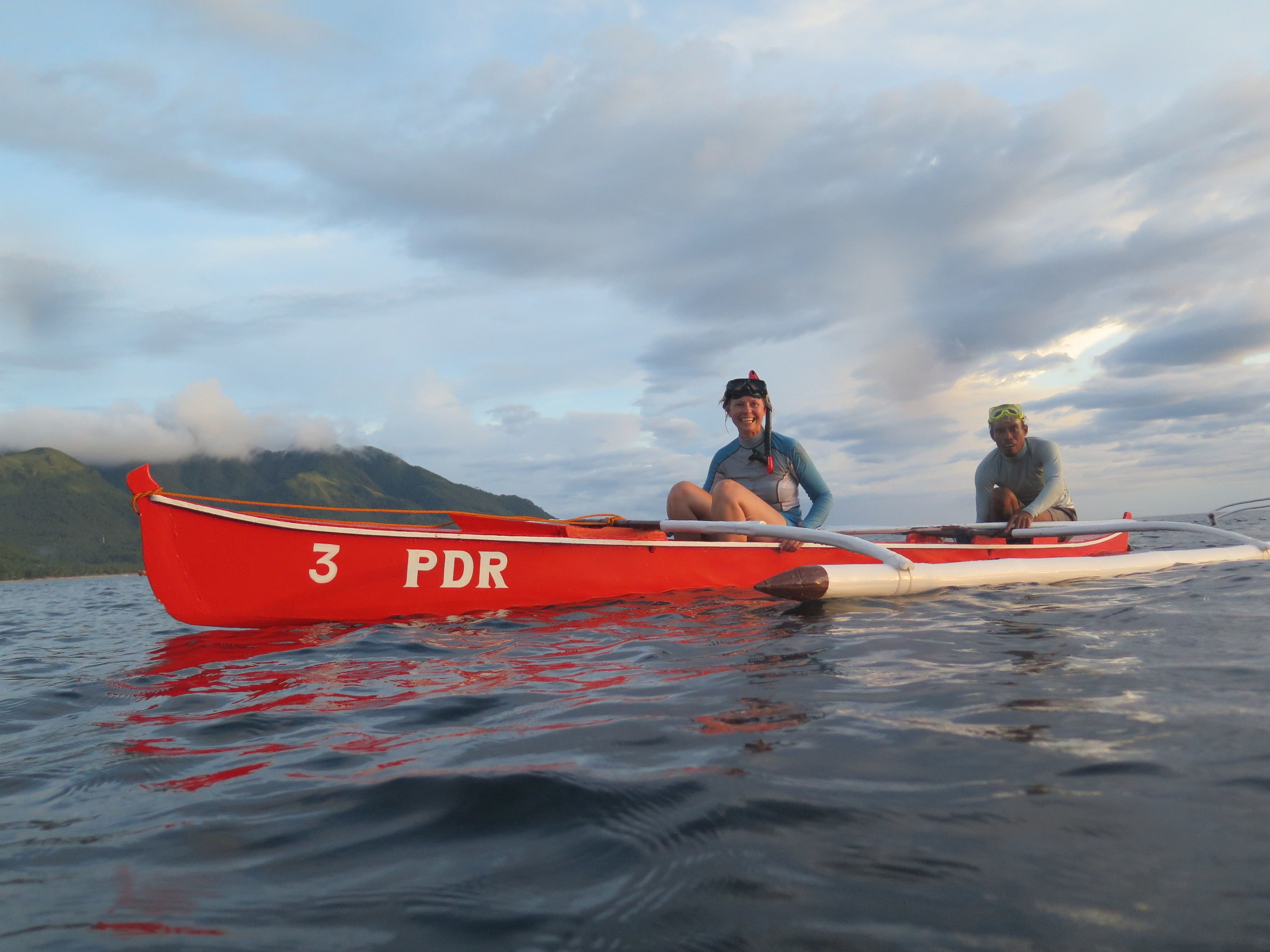 Deux personnes dans un bateau rouge portant le numéro 3 PDR, sur une étendue d’eau calme, avec des montagnes en arrière-plan.