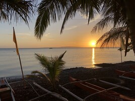 Escena de playa con barcos, una bandera y palmeras al atardecer.