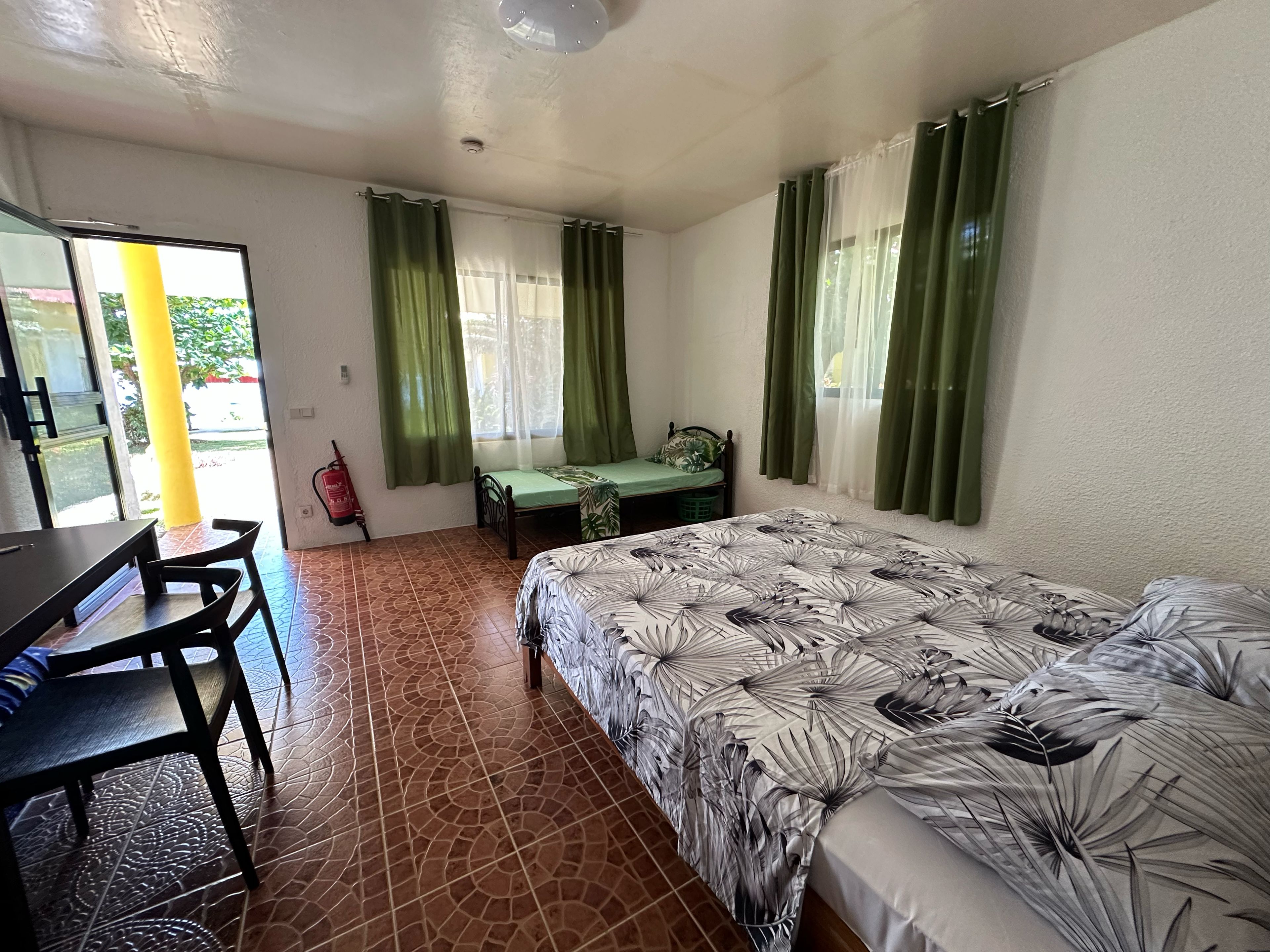 A room with a bed, a desk, and a chair. There is a window with curtains, a fire extinguisher, and a door.