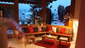 Ein gemütliches Wohnzimmer mit einem holzgestellten Ecksofa, roten und braunen Kissen sowie einem roten Teppich. Zwei Holztische und eine Stehlampe sind ebenfalls vorhanden. Ein großes Fenster bietet Blick auf eine verschneite Landschaft.