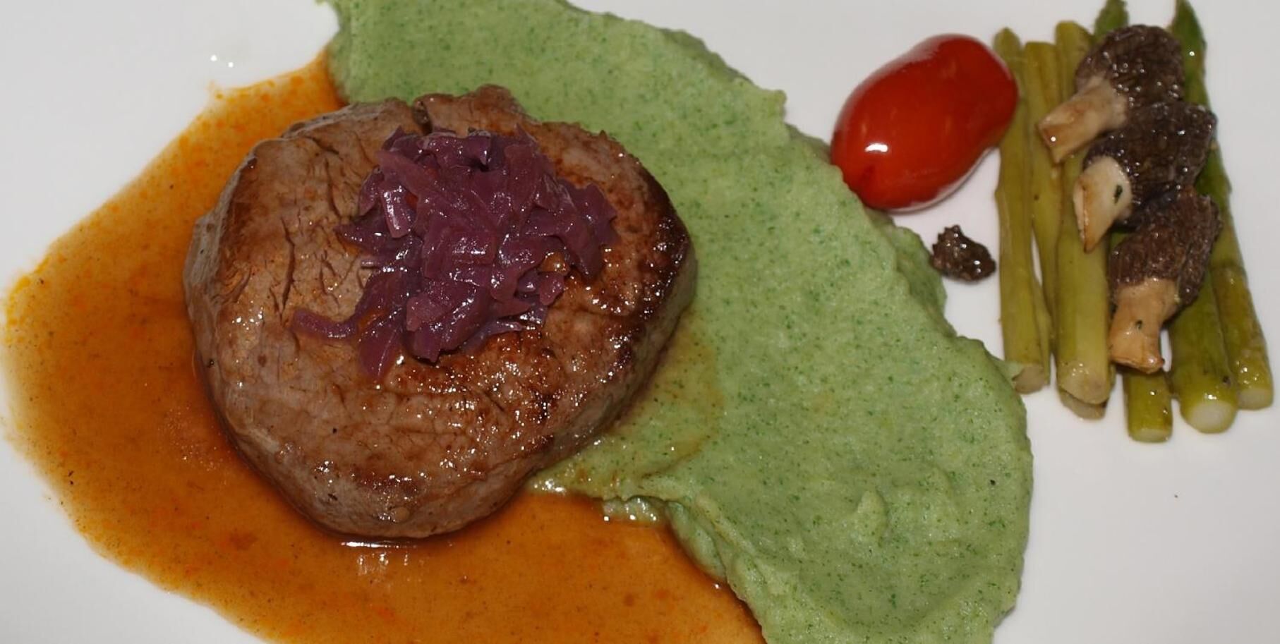 A plate with a steak topped with red onions, a green sauce, asparagus, and a cherry tomato.