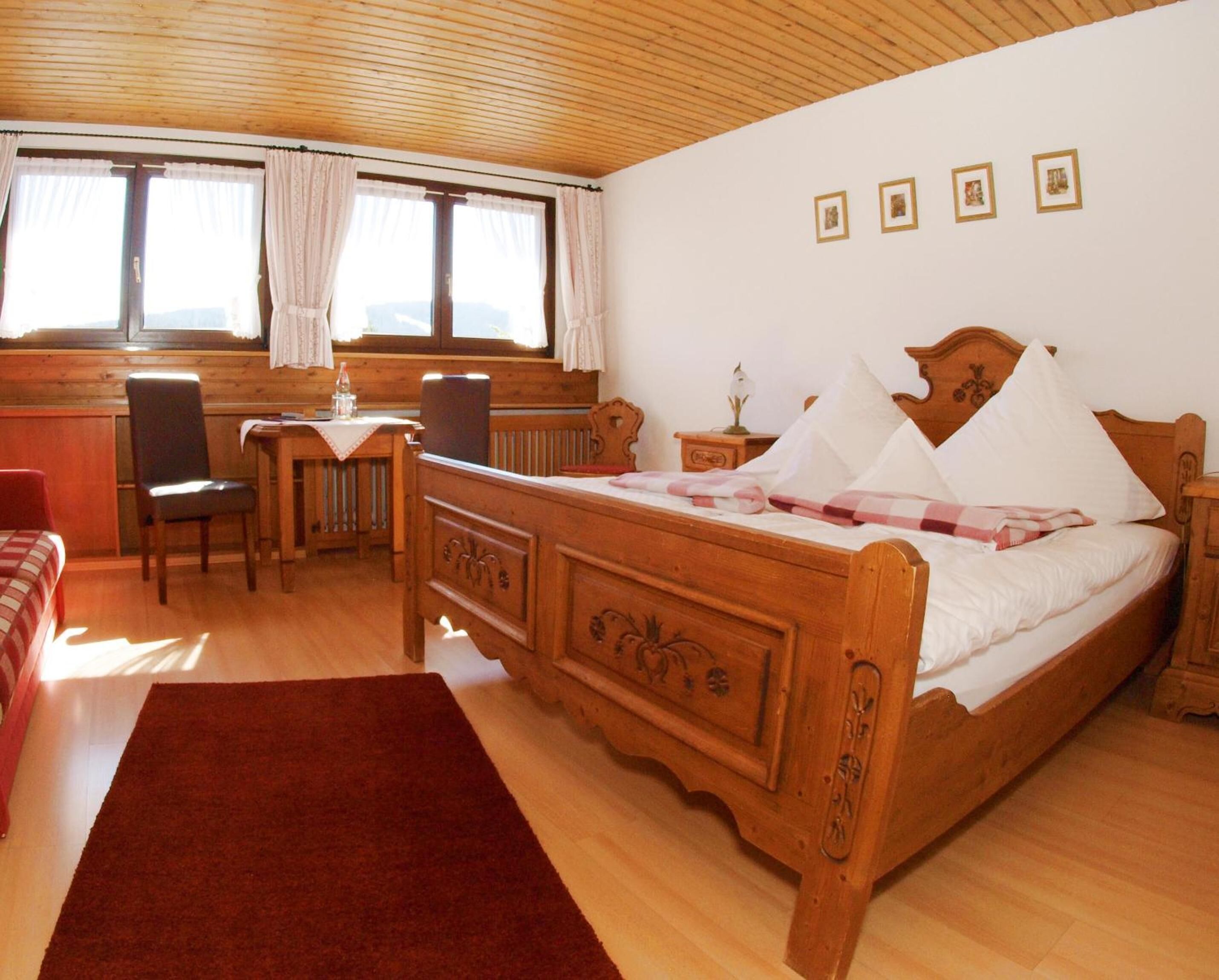 Ein hölzernes Bett mit einer rot-weißen Karodecke, ein Schreibtisch aus Holz mit einem Stuhl und ein Fenster mit Vorhängen.