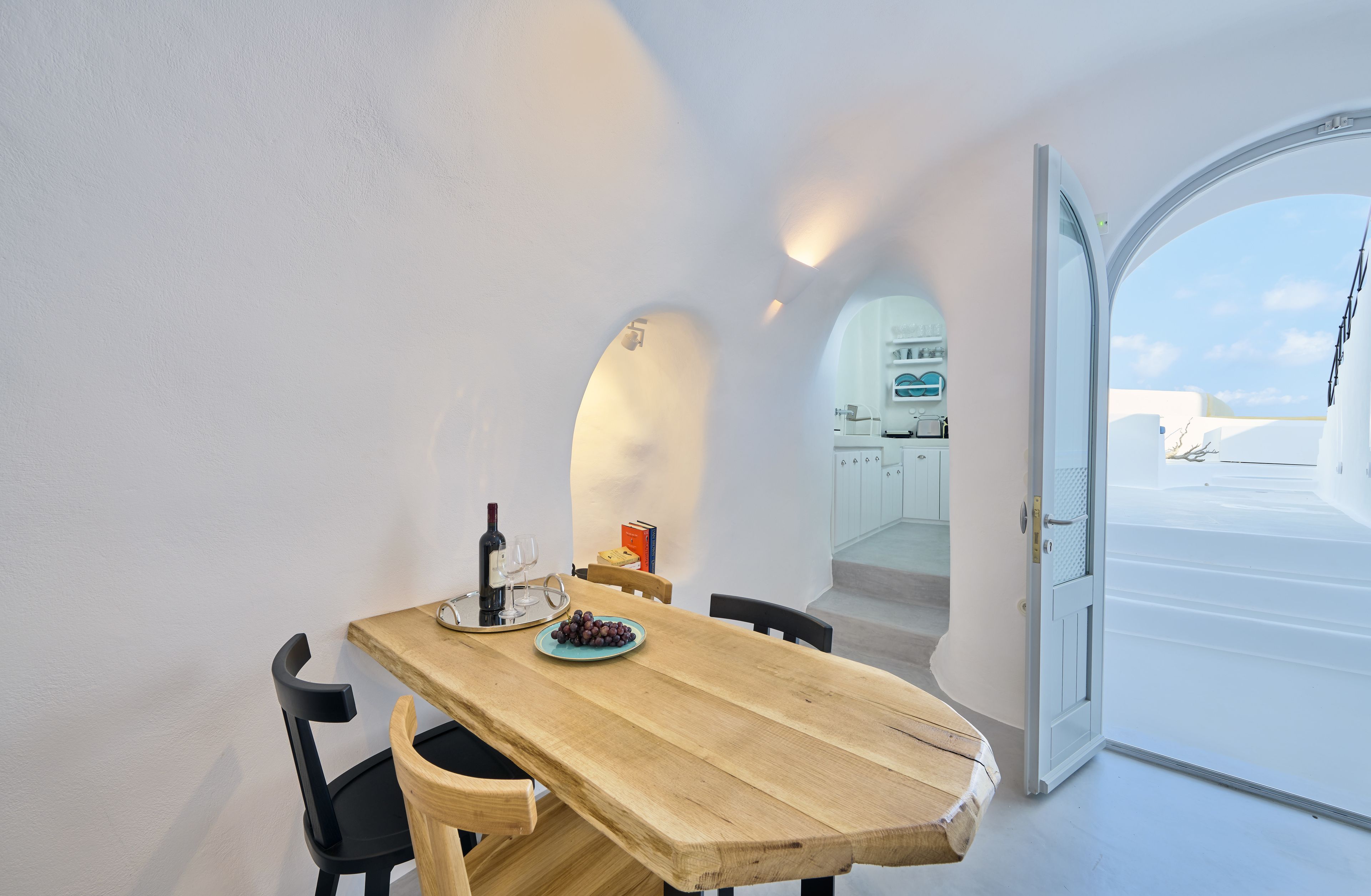 Un comedor con una mesa de madera, sillas negras, un plato de uvas, una botella de vino y copas. Hay una puerta blanca que da a un balcón con vistas al cielo y edificios.