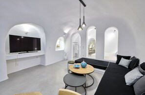 A modern living room with a large flat-screen TV, a black sofa, a round wooden table, and a small round table with a bowl and vase.