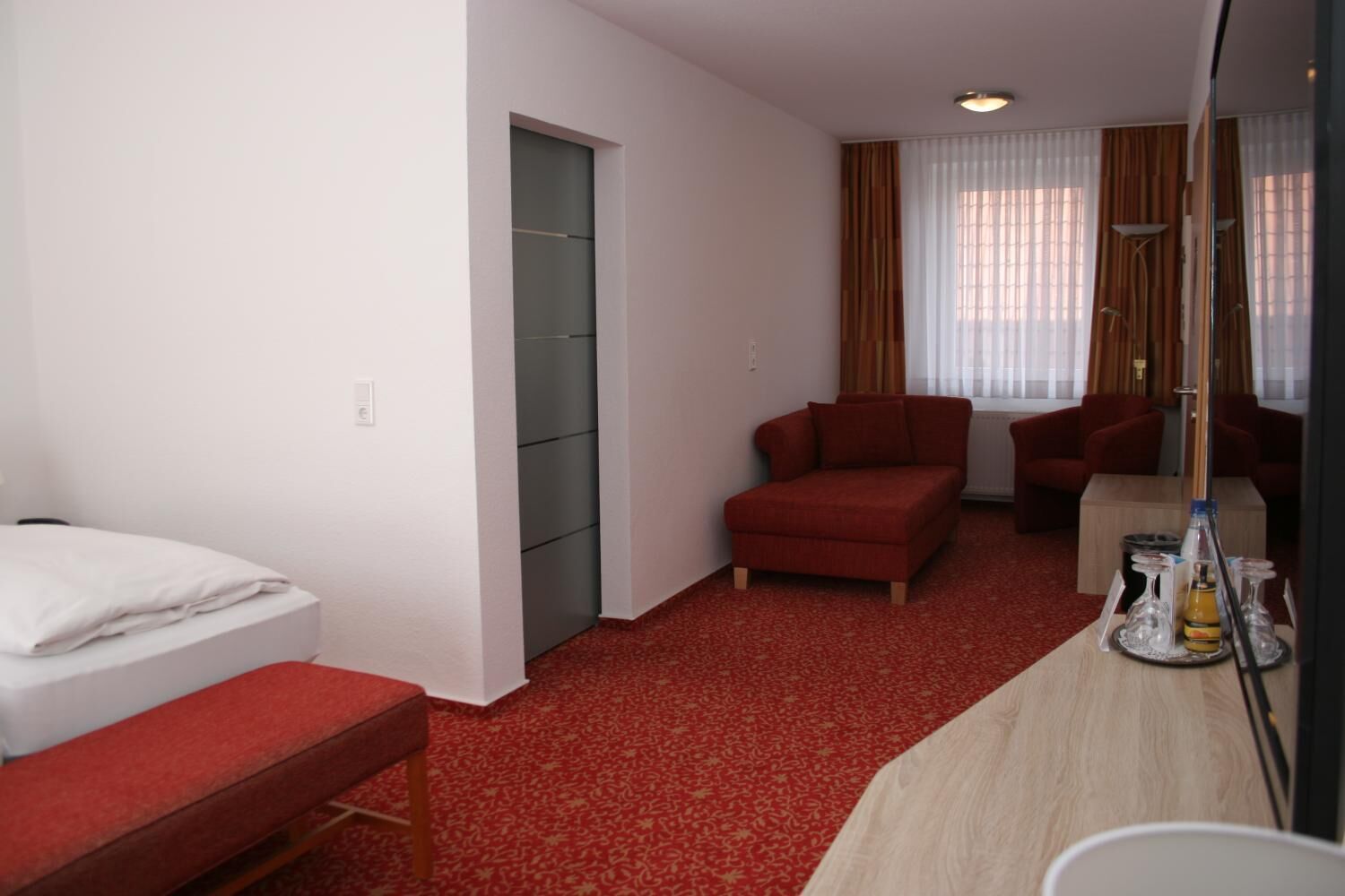 A hotel room with a bed, a red sofa, a desk with a mirror, and a window with curtains.