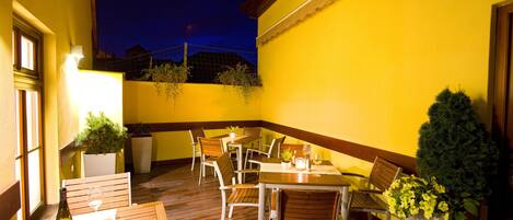 An outdoor patio with wooden tables and chairs, potted plants, and a bottle of wine.