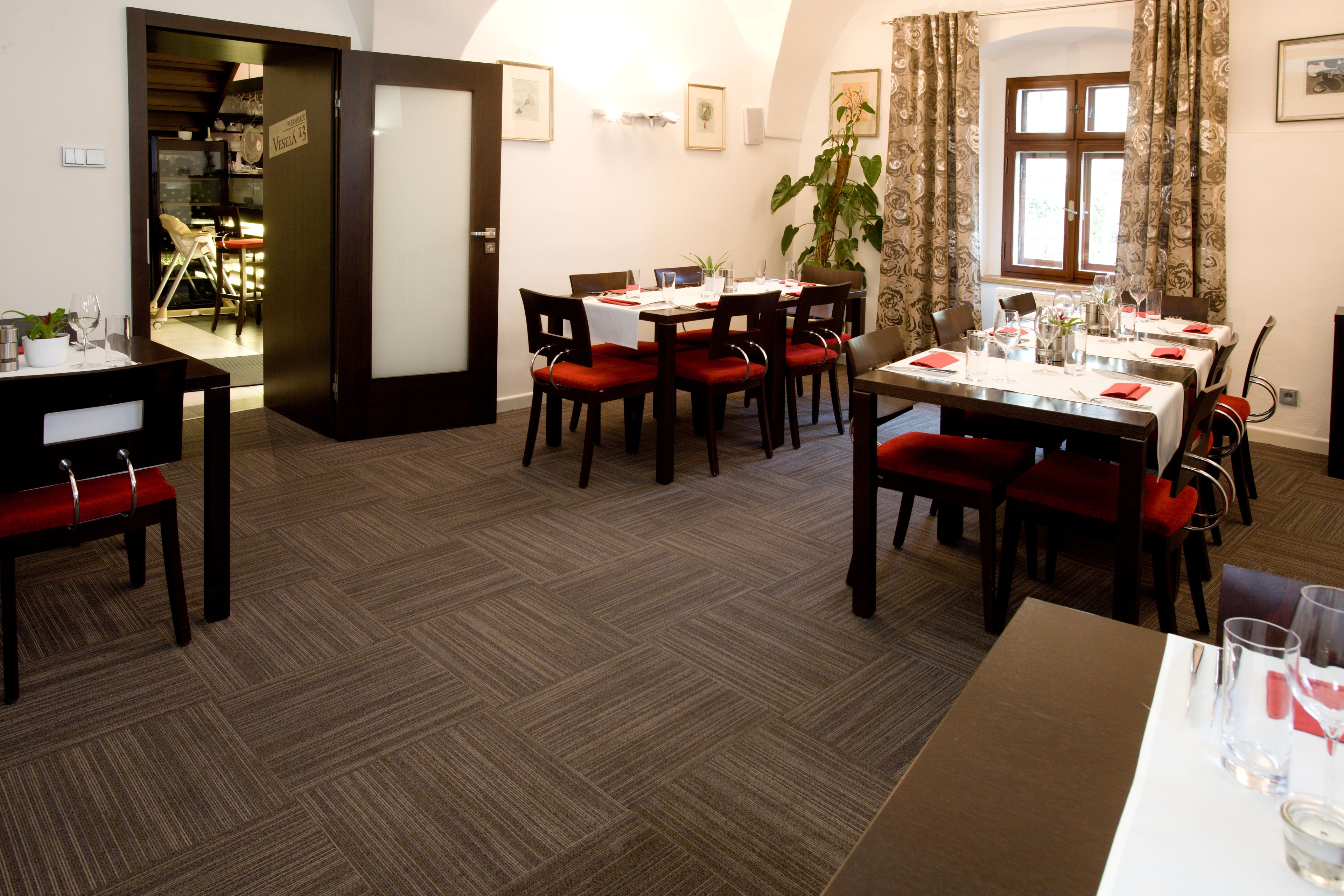 A dining area with tables set for four, red upholstered chairs, and a large potted plant.