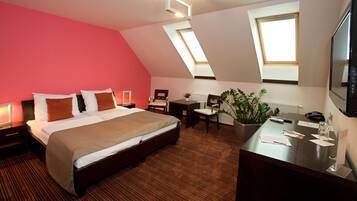 Ein Hotelzimmer mit einem großen Bett, einem Schreibtisch, zwei Stühlen, einer Zimmerpflanze und einem Fernseher.