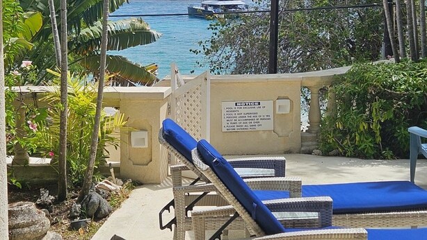 A covered pool area with lounge chairs, a notice board, and a view of the ocean with boats.