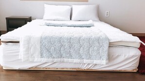 A neatly made bed with white bedding and a light blue throw blanket, a small bedside table, and a window with a view of buildings. - Leeds Pension (Seogwipo)