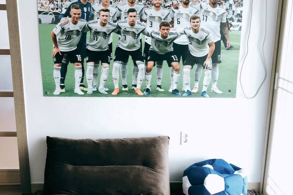 A room with a brown sofa, a framed soccer team photo on the wall, and a blue and white soccer ball on the floor.