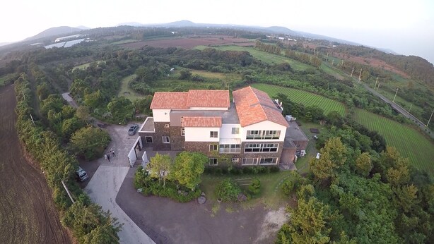 A large, two-story house with a red-tiled roof, surrounded by lush greenery and fields. - Leeds Pension (Seogwipo)