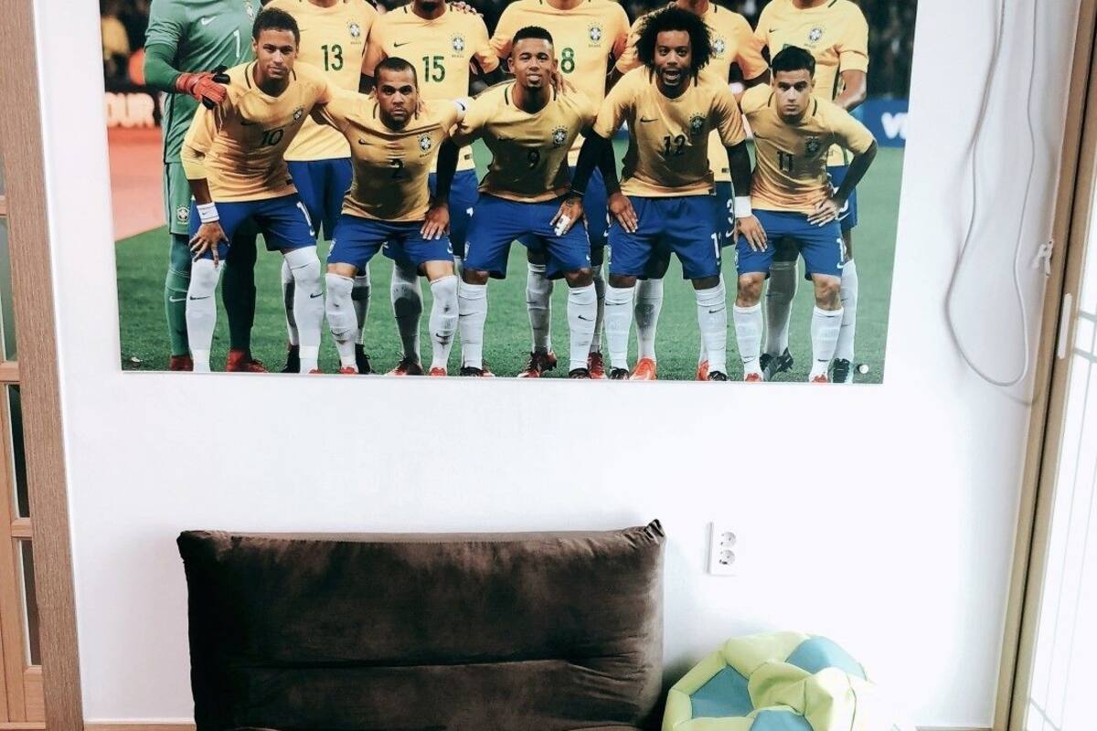 A room with a brown sofa, a wall-mounted television displaying a soccer team, and a window with blinds.