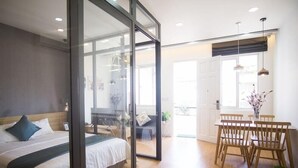 A modern hotel room with a large bed, a dining area with wooden chairs, and a glass partition.