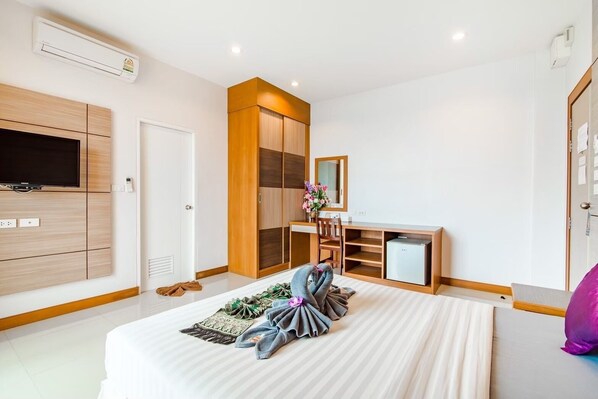A modern bedroom with a large bed, a TV mounted on the wall, a wardrobe, a desk with a mirror, and a small fridge.