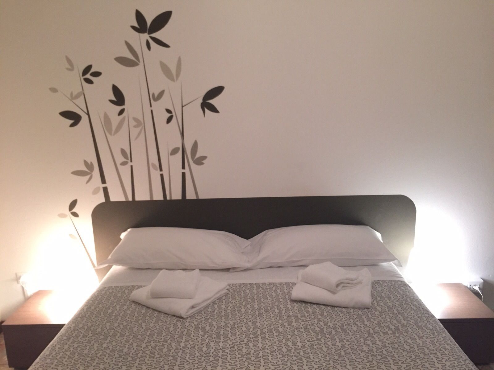 Una cama bien tendida con sábanas y toallas blancas, un cabecero oscuro y una decoración de hojas estilizadas.